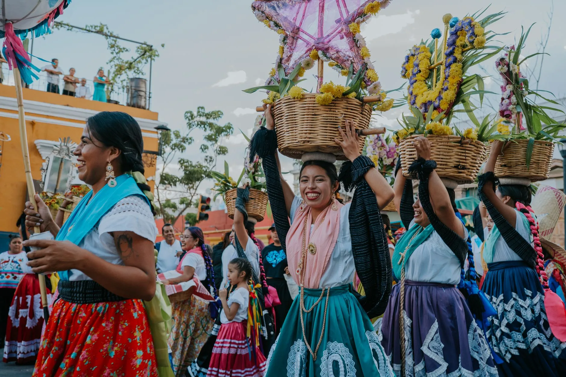 Femmes en costumes traditionnels colorés défilant avec des paniers de fleurs sur la tête lors d'une fête, sourires et foule autour